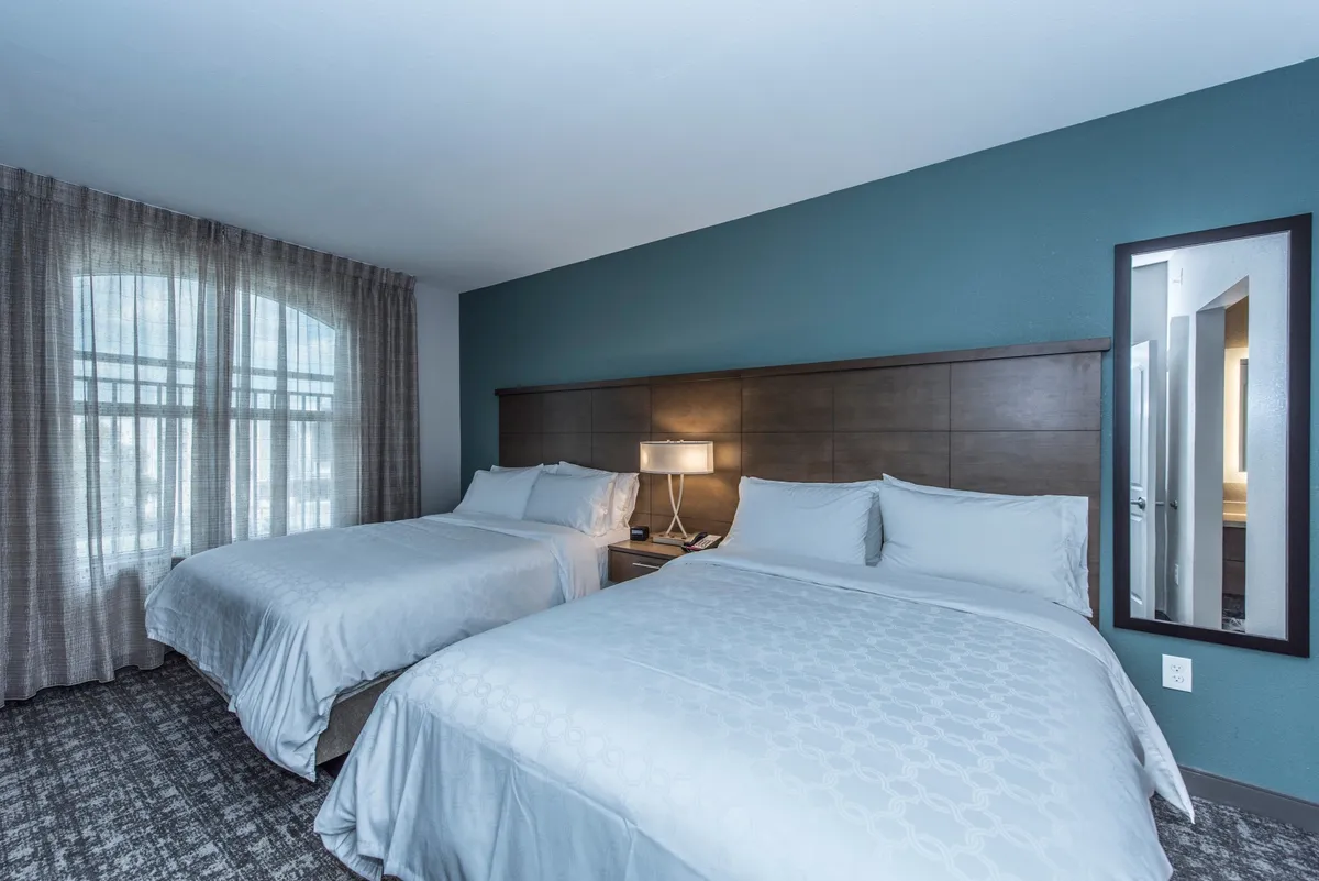 A tranquil bedroom with two beds, a nightstand and lamp, and a large window letting in natural light. The arrangement is practical for families or groups, providing both personal space and opportunities for late-night conversation.