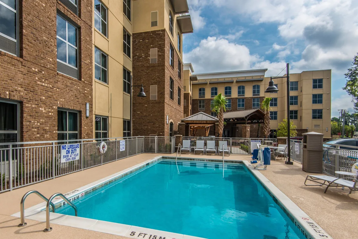 An inviting outdoor swimming pool is surrounded by several lounge chairs and bordered by well-maintained landscaping and the property’s buildings. The area is bathed in natural sunlight, offering a relaxing and communal environment ideal for summer leisure.