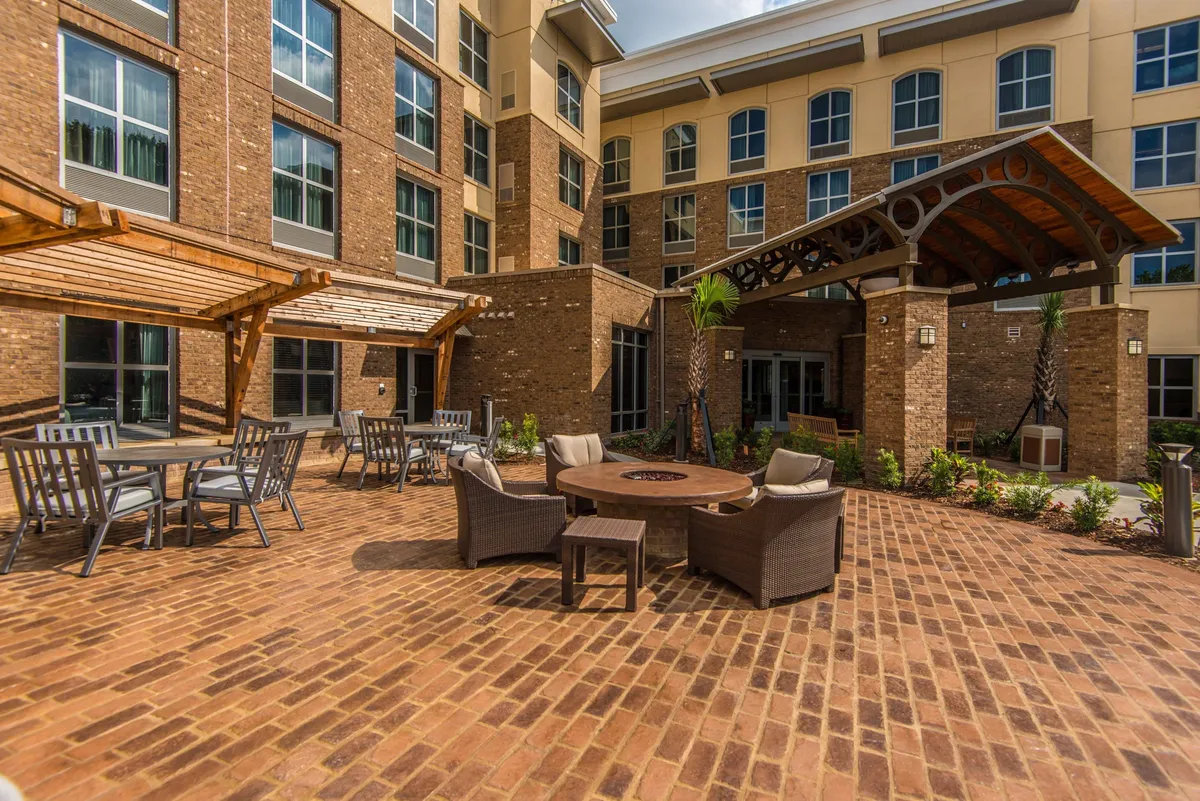 An inviting fire pit area is at the center of this outdoor scene, with comfortable seating and palm trees adding to the relaxed, resort feel. The brick patio signals a space for socializing, storytelling, or unwinding after a day of exploring nearby beaches, parks, and historic sites.