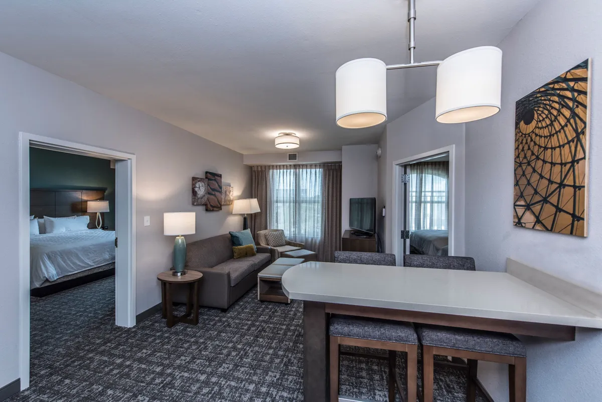 A bright, inviting condo interior featuring a plush sofa perfect for movie nights, a modern dining table set for family meals or morning coffee, stylish artwork on the wall, and a glimpse into a cozy bedroom beyond.