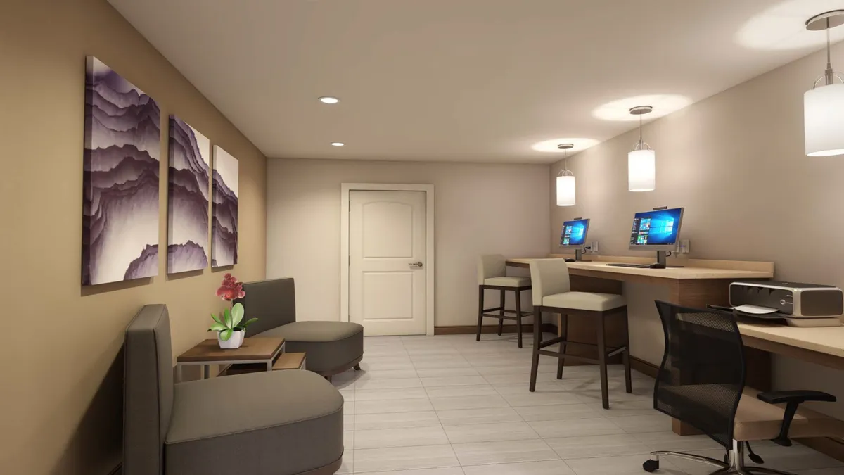 This image captures a light-filled business center area outfitted with computers on desks, comfortable seating areas, and inspiring mountain-themed wall art. The setting feels both productive and relaxed, with a layout designed for collaborative work or focused solo productivity.