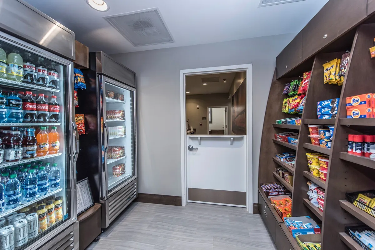 A brightly lit snack and beverage area with a refrigerated section and shelves of grab-and-go items, inviting guests to pick up refreshments as they come and go.