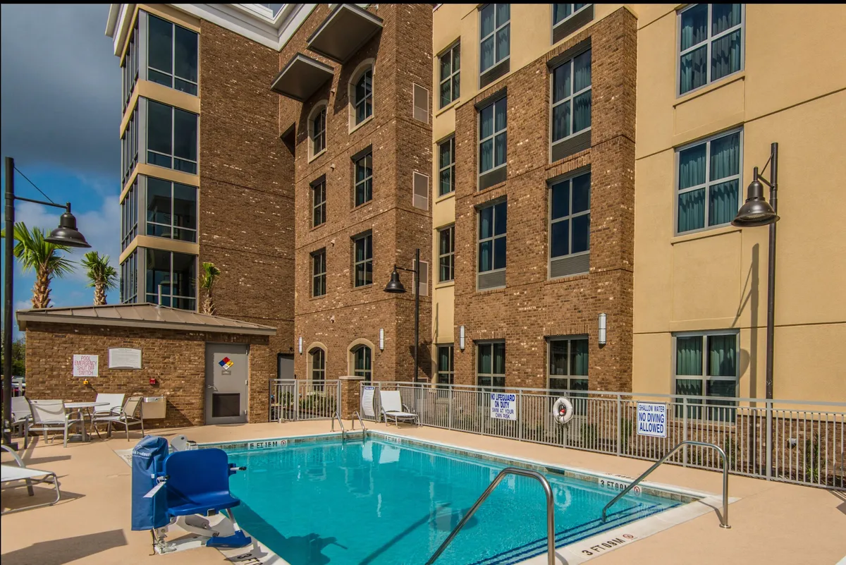 A sparkling swimming pool is set against palm trees and surrounding buildings, with multiple lounge chairs ready for relaxation. The relaxed atmosphere is complemented by the inviting blue water and ample space for sunbathing or socializing poolside.