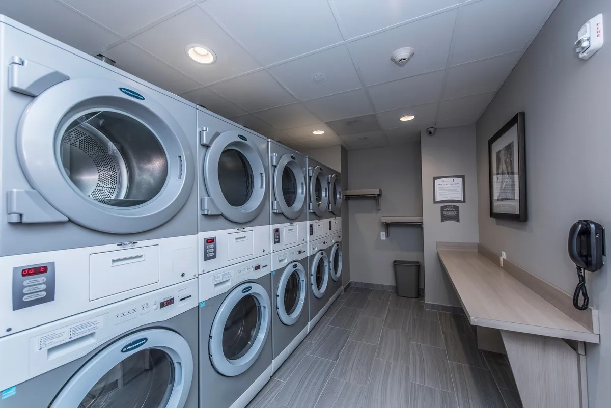 A dedicated laundry room with a row of modern washing machines and dryers, a convenient folding counter, and bright artwork on the wall. The space is clean, organized, and practical, showcasing essential amenities for longer stays.