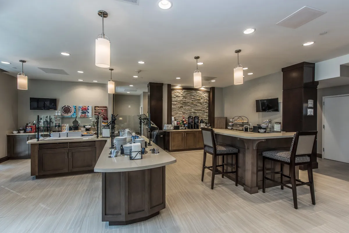 The scene captures a bright breakfast area with a central bar counter, beverage station, and ample seating. The design is modern and welcoming, ideal for starting the day with complimentary breakfast or informal group gatherings.